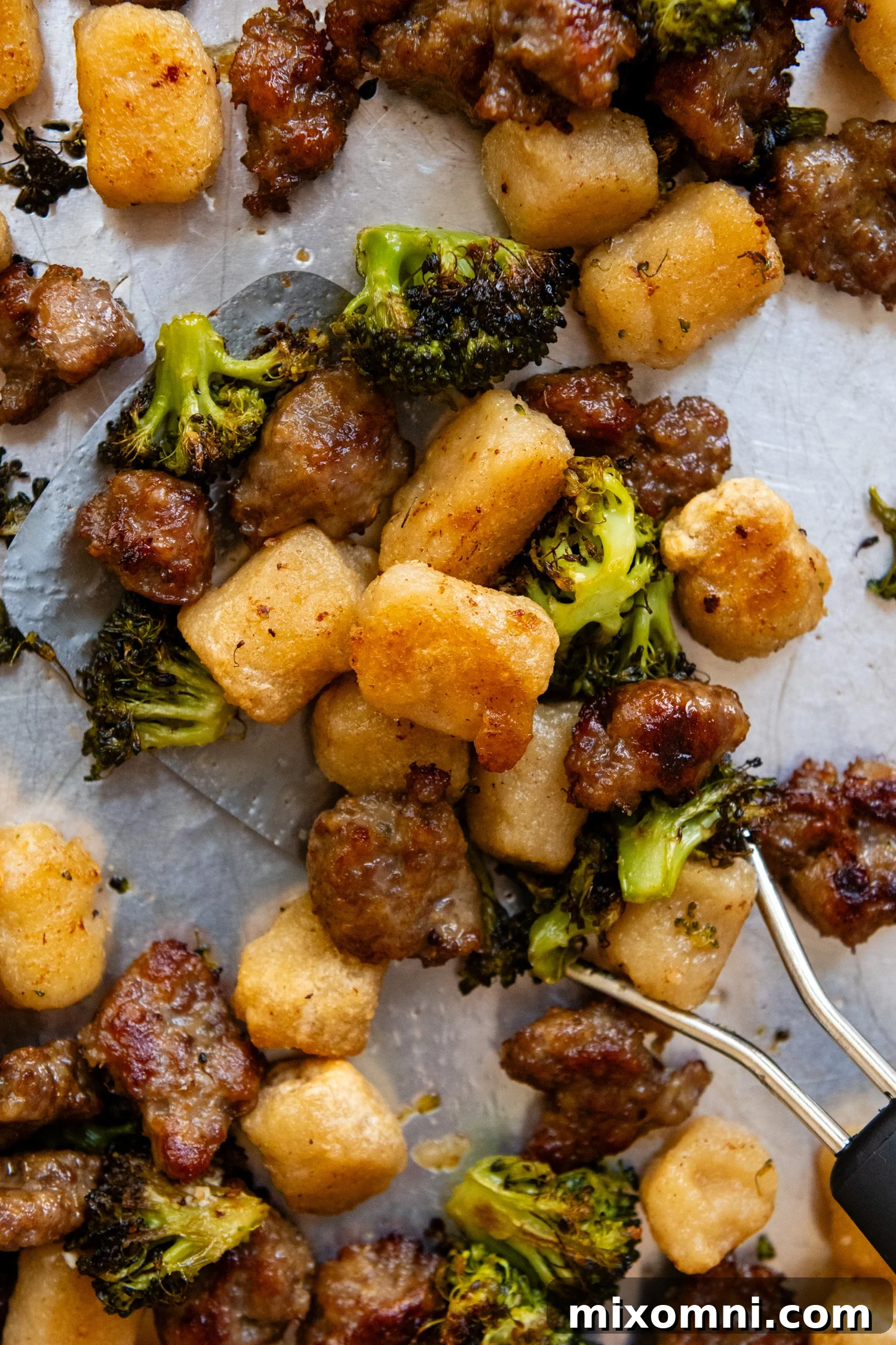 Close up of perfectly cooked crispy gnocchi, roasted broccoli florets, and browned Italian sausage pieces on a metal spatula, ready to be served.