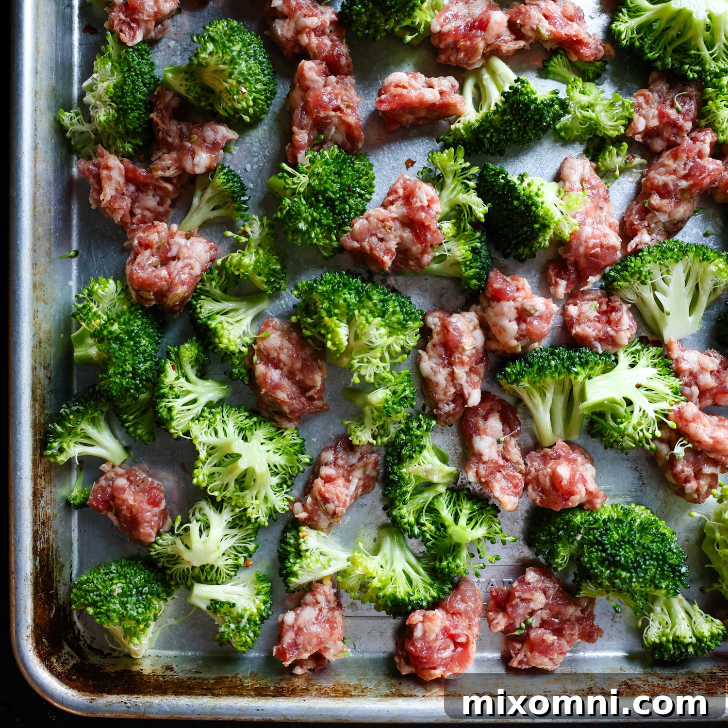 Raw broccoli florets and crumbled Italian sausage pieces arranged on a sheet pan, seasoned with olive oil and salt, ready for roasting.