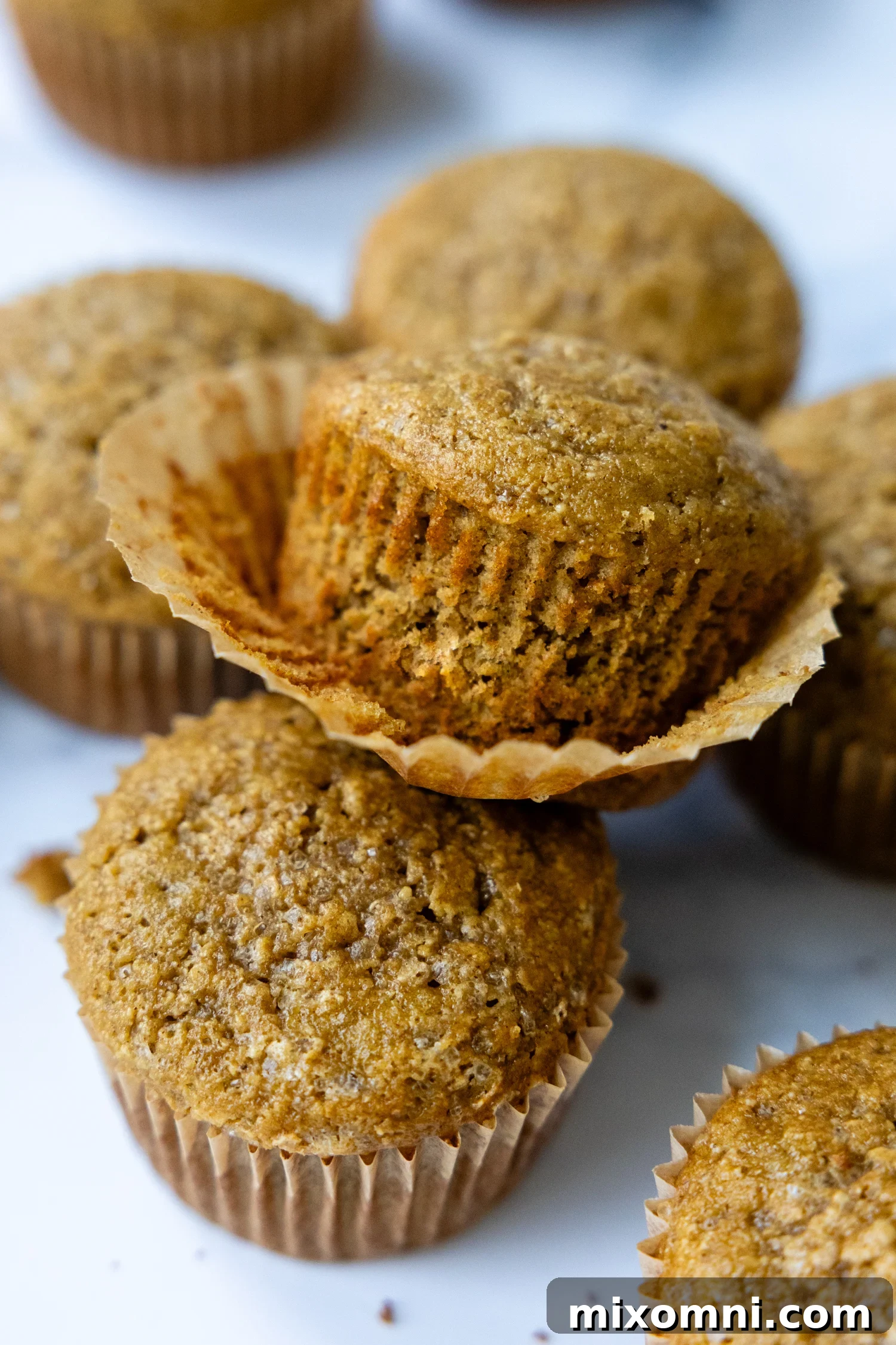 A stack of golden-brown oat flour banana muffins, with one muffin partially unwrapped to show its tender, moist texture.