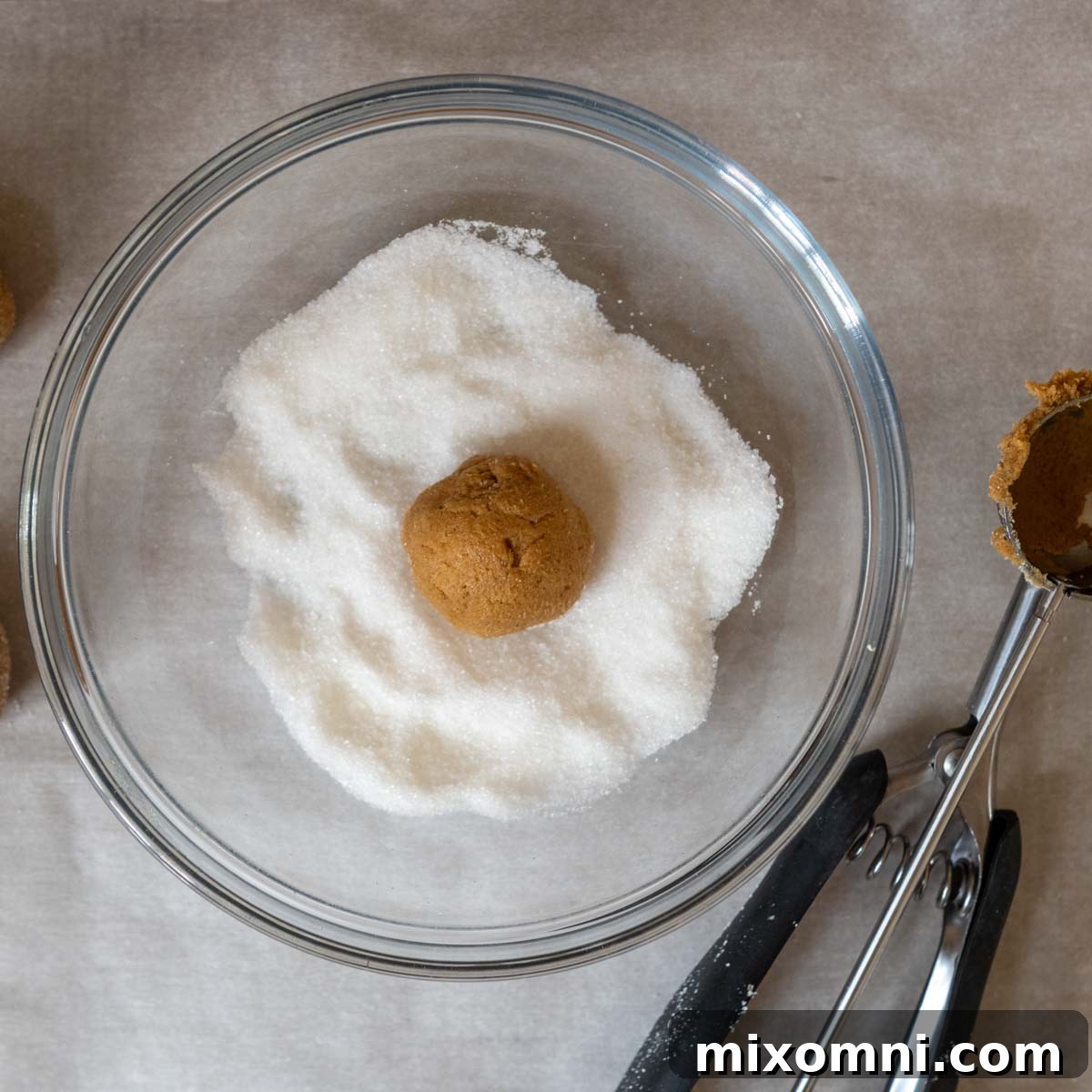 A perfectly portioned gluten-free ginger snap cookie dough ball being rolled in a bowl of sparkling granulated sugar, preparing it for baking and a delightful crinkled finish.