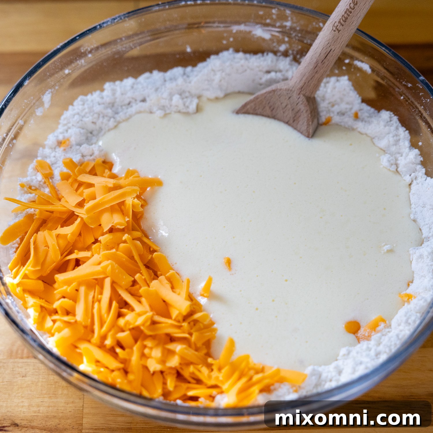 A bowl of dry biscuit ingredients mixed with shredded cheese, and a separate bowl of milk and other wet ingredients.