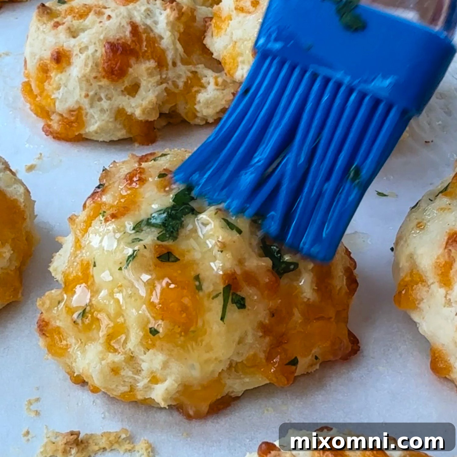 A blue basting brush spreading melted garlic butter on hot, cooked biscuits.