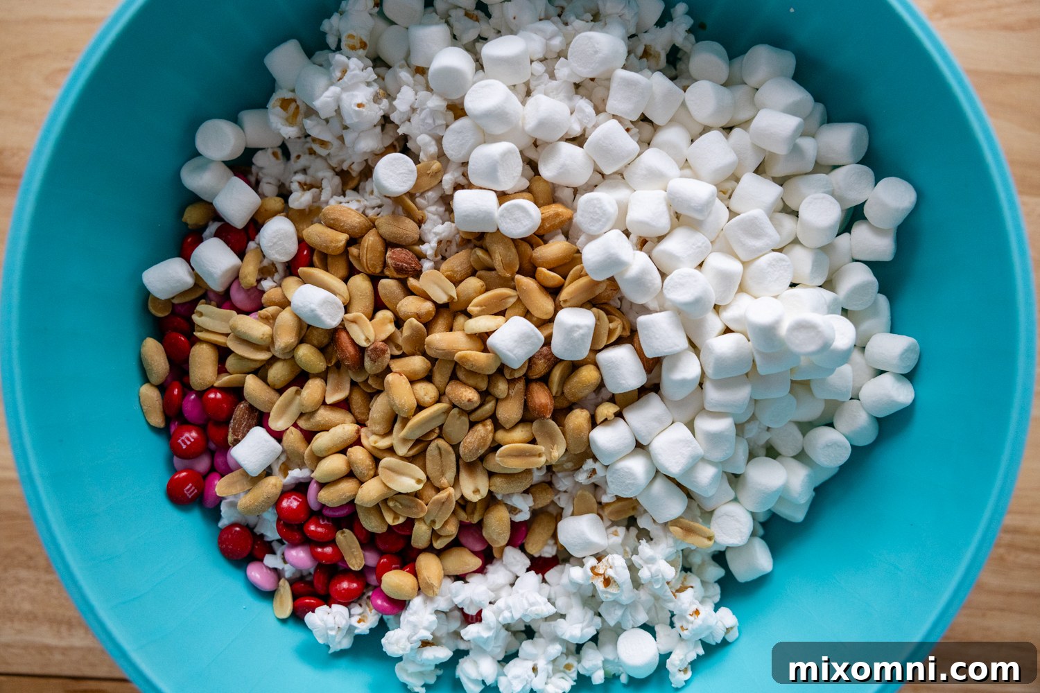 Peanuts, marshmallows, M&M's, and popcorn are mixed together in a large teal bowl, ready for coating.