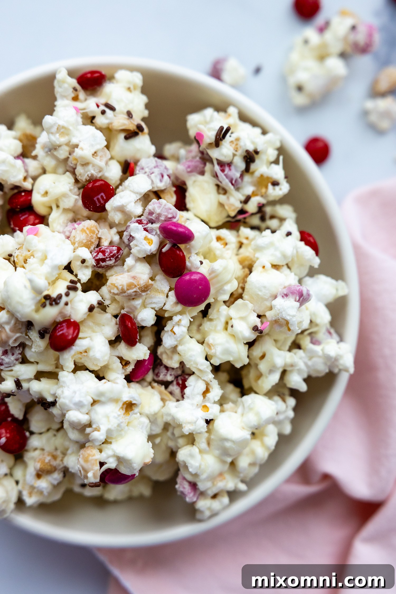 A bowl filled with white chocolate popcorn mix, accompanied by a pink towel on a white surface.