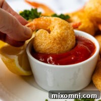 A hand dipping a crispy gluten-free fried shrimp into a white dish of shrimp sauce.