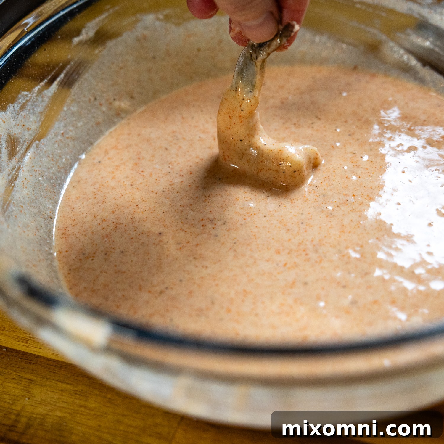 Raw shrimp being dipped into the prepared gluten-free batter, held by its tail.