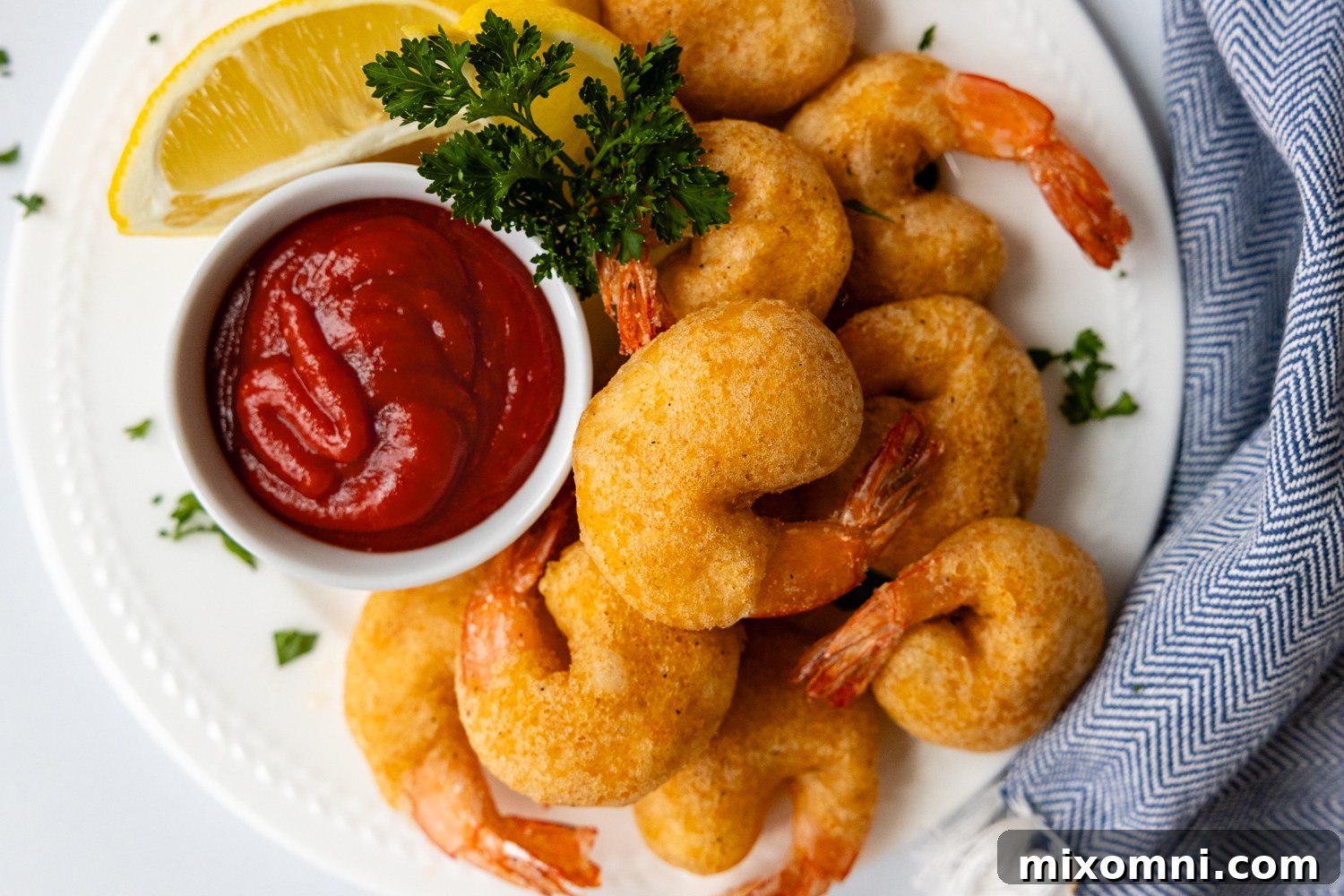 Overhead shot of a white plate piled high with golden, crispy fried shrimp, accompanied by a small dish of shrimp sauce and lemon wedges.
