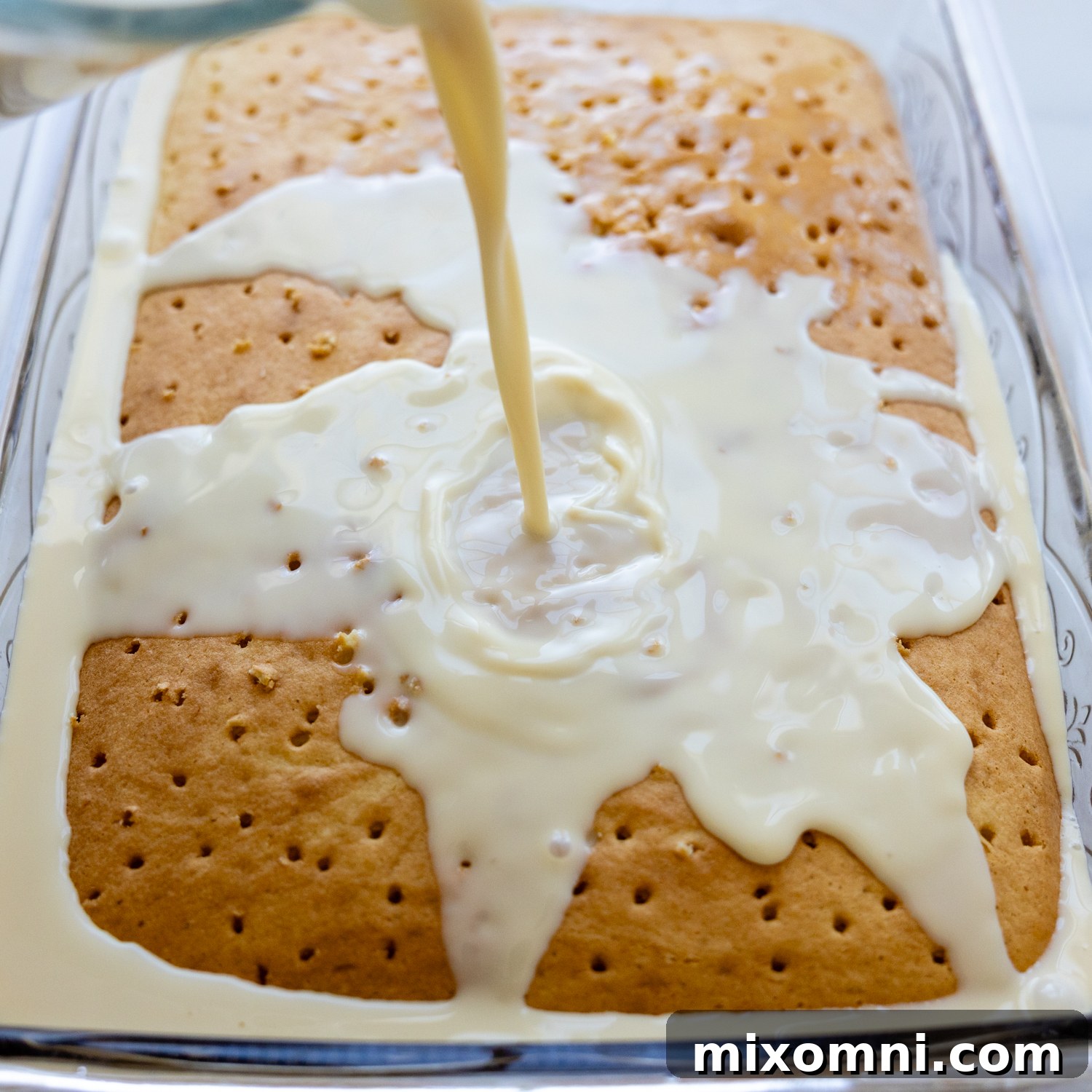 The baked cake with holes poked into it, slowly absorbing the rich three-milk soak being poured over its surface.