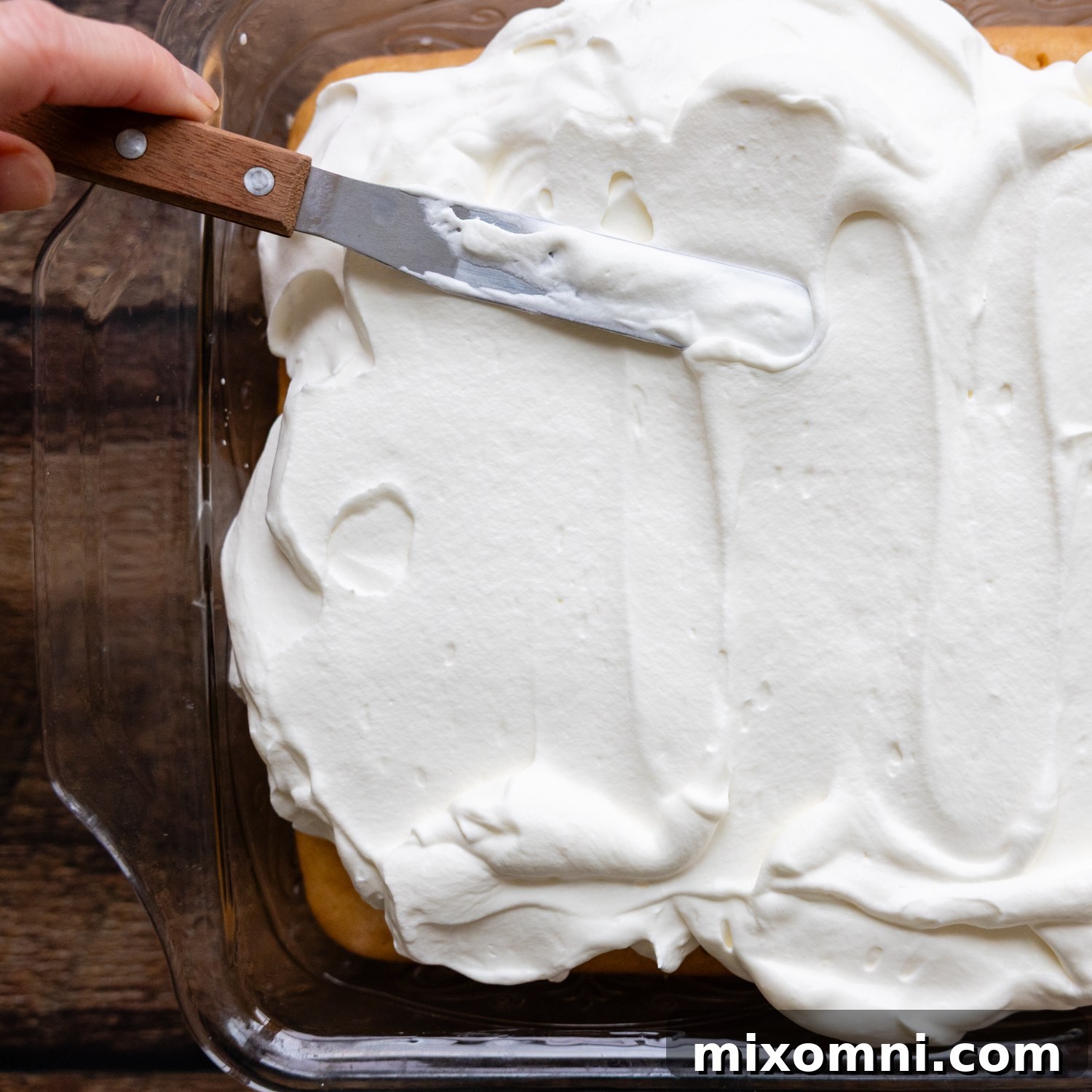 A gluten-free tres leches cake being generously frosted with freshly made whipped cream.