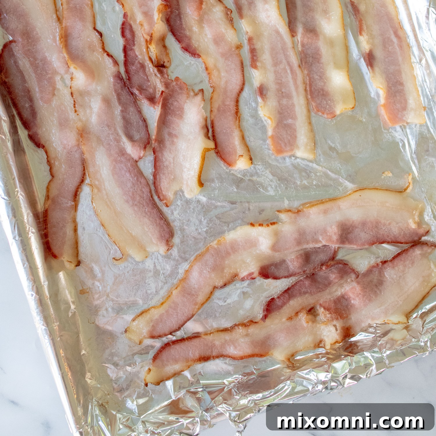 Partially baked bacon strips laid out on a foil-lined baking sheet, golden and crisping.