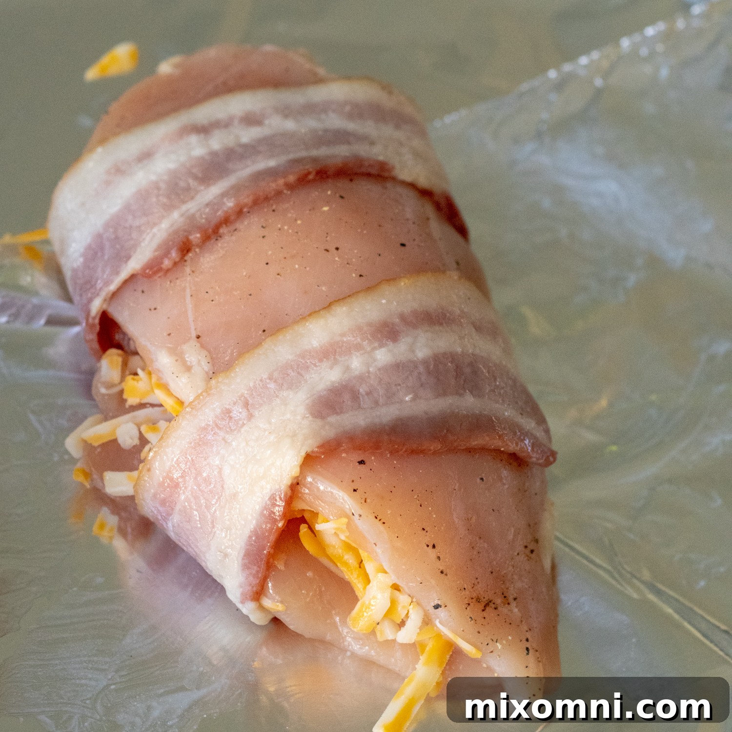 Partially cooked bacon strips wrapped tightly around a cheese-stuffed chicken breast on a baking sheet.