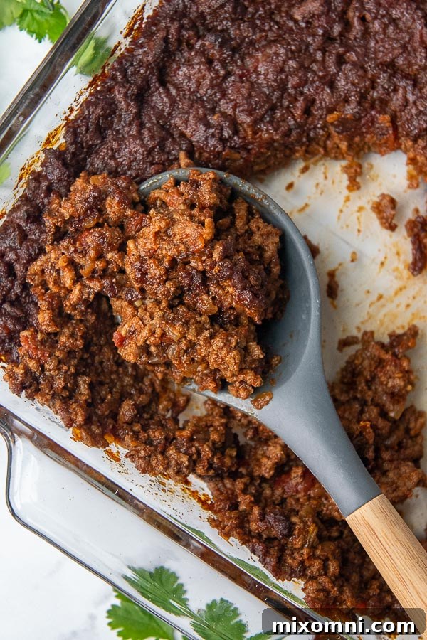 Overhead shot of a wooden spoon scooping up spoonfuls of rich, spicy, and sweet taco meat from a baking dish.