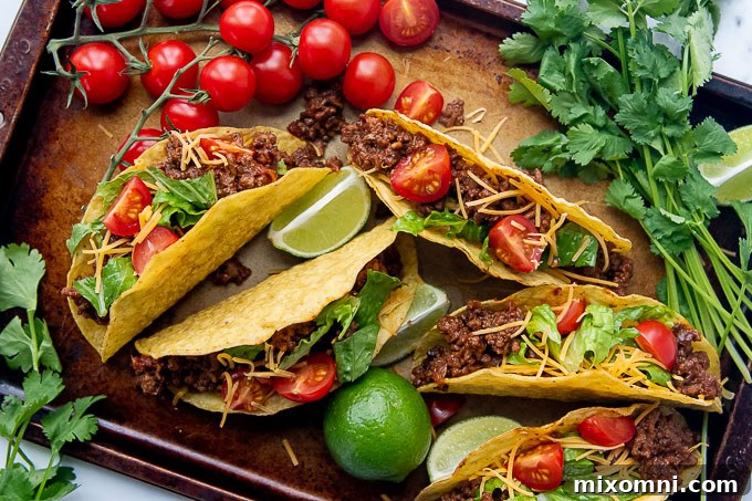 Overhead shot of vibrant spicy ground beef tacos arranged on a baking sheet, surrounded by various fresh toppings like cheese, lettuce, and cilantro.