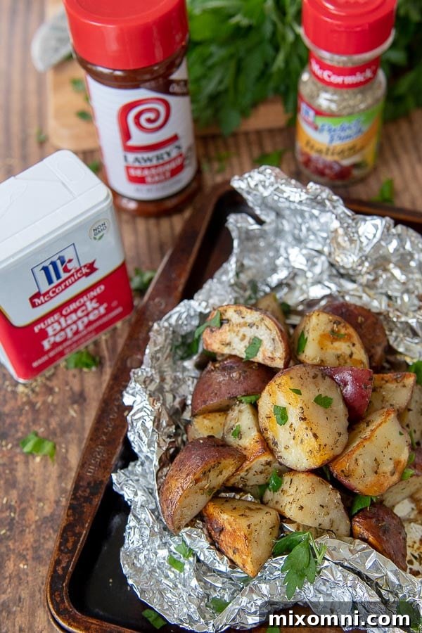 A foil packet of perfectly grilled potatoes rests on a baking sheet, surrounded by the fresh herbs and spices used to create its delicious flavor.