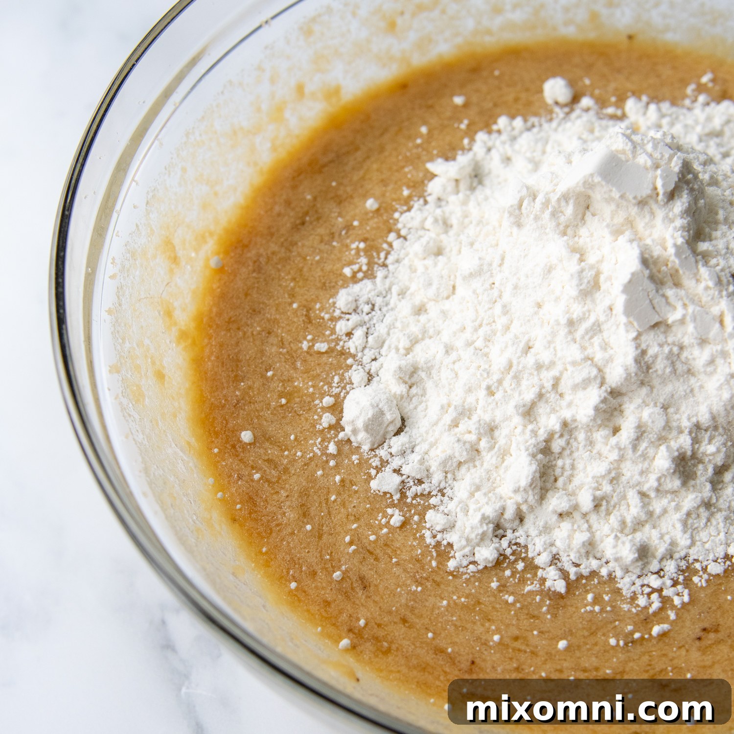 Gluten-free flour being gently mixed into the banana batter in a large bowl, creating a smooth consistency.