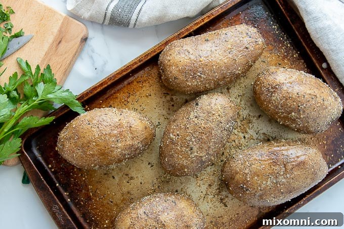 A baking sheet of baked potatoes with seasoned crispy skins and sprigs of parsley laying next to it.