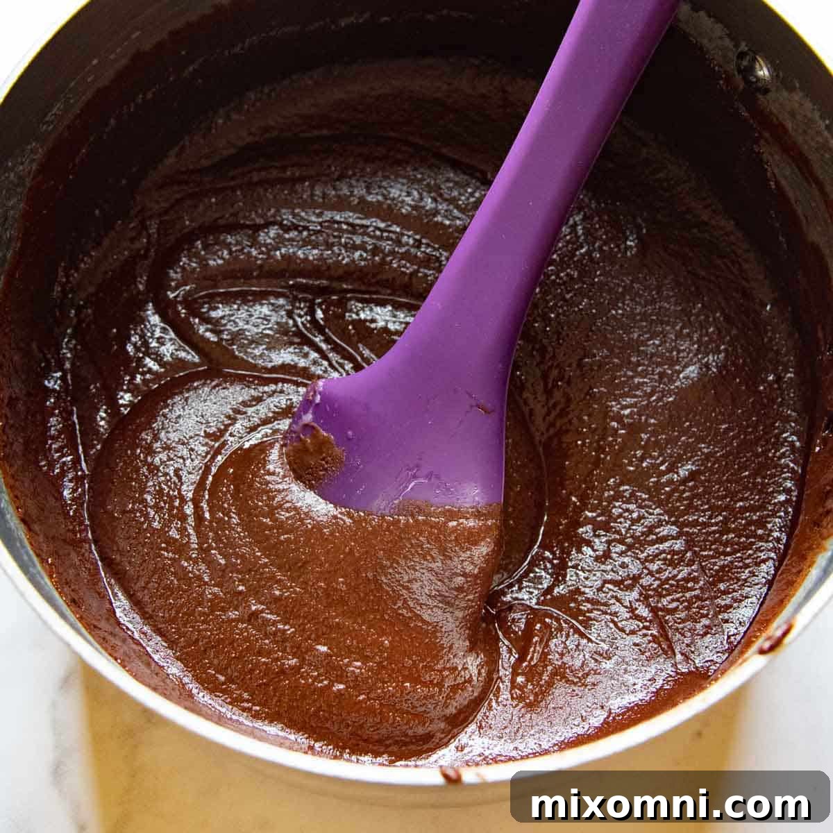 A purple spatula stirring a rich, melted chocolate mixture in a saucepan, creating a smooth and glossy base for the brownies.
