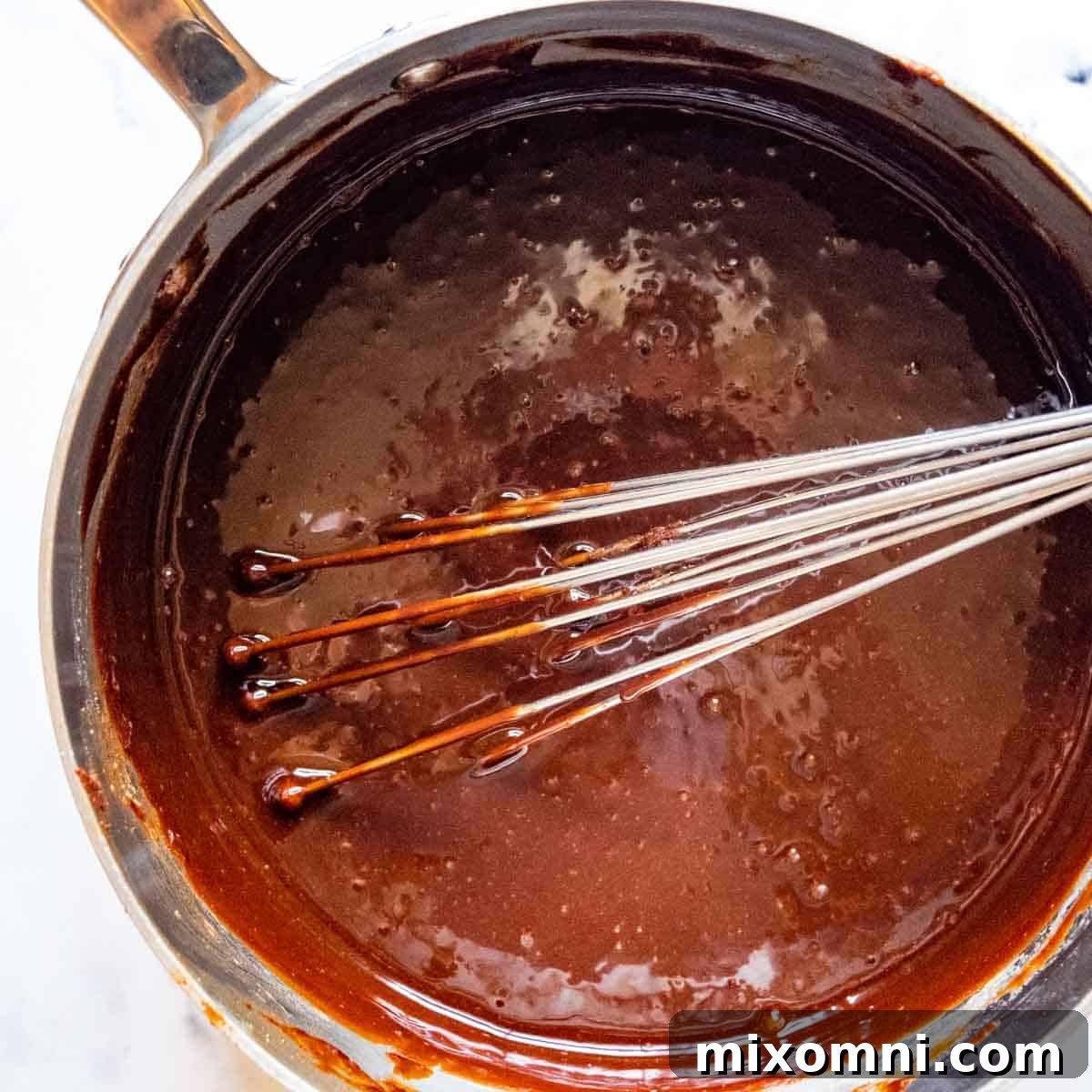 A whisk vigorously mixing eggs and vanilla extract into the cooled chocolate mixture in a saucepan, aiming for a glossy finish.