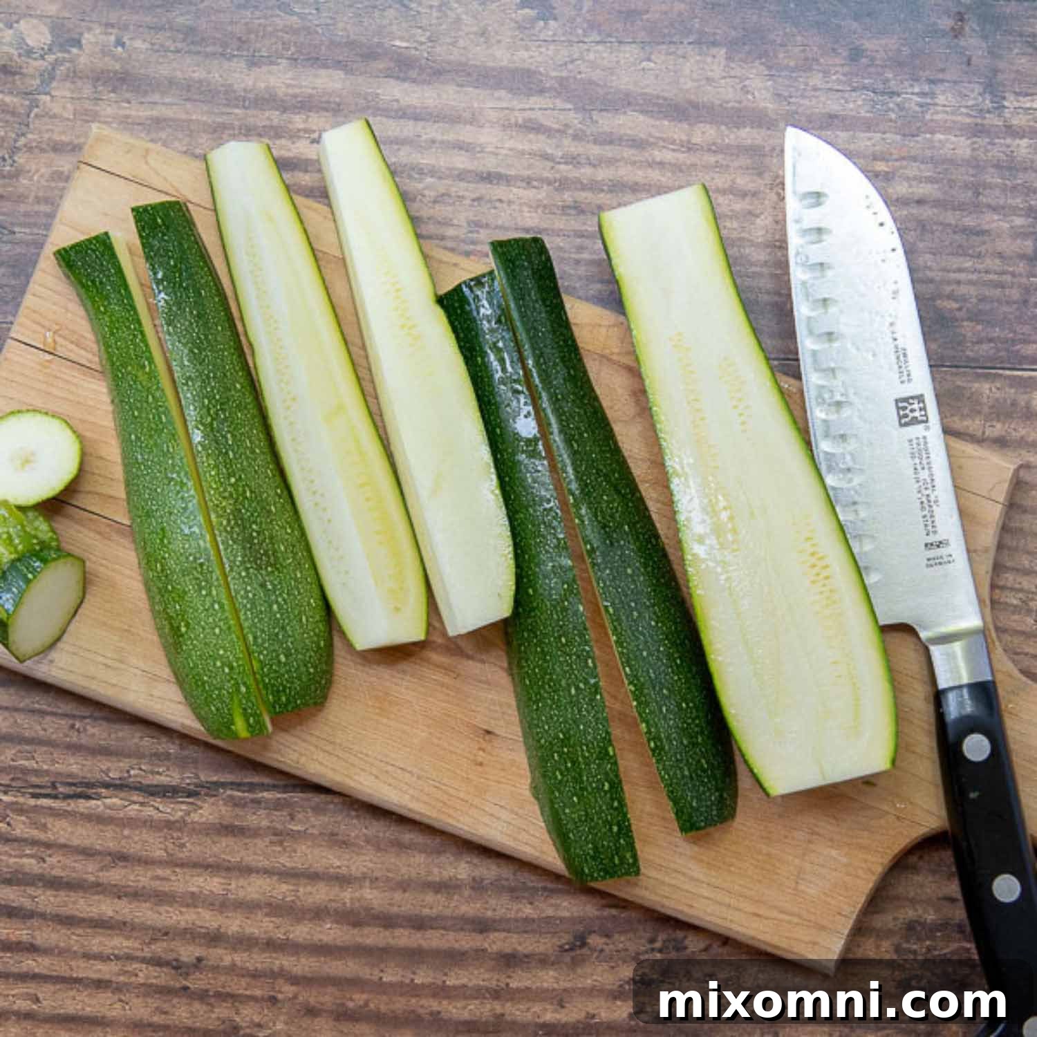 Zucchini being sliced lengthwise into spears on a wooden cutting board with a chef's knife beside it.
