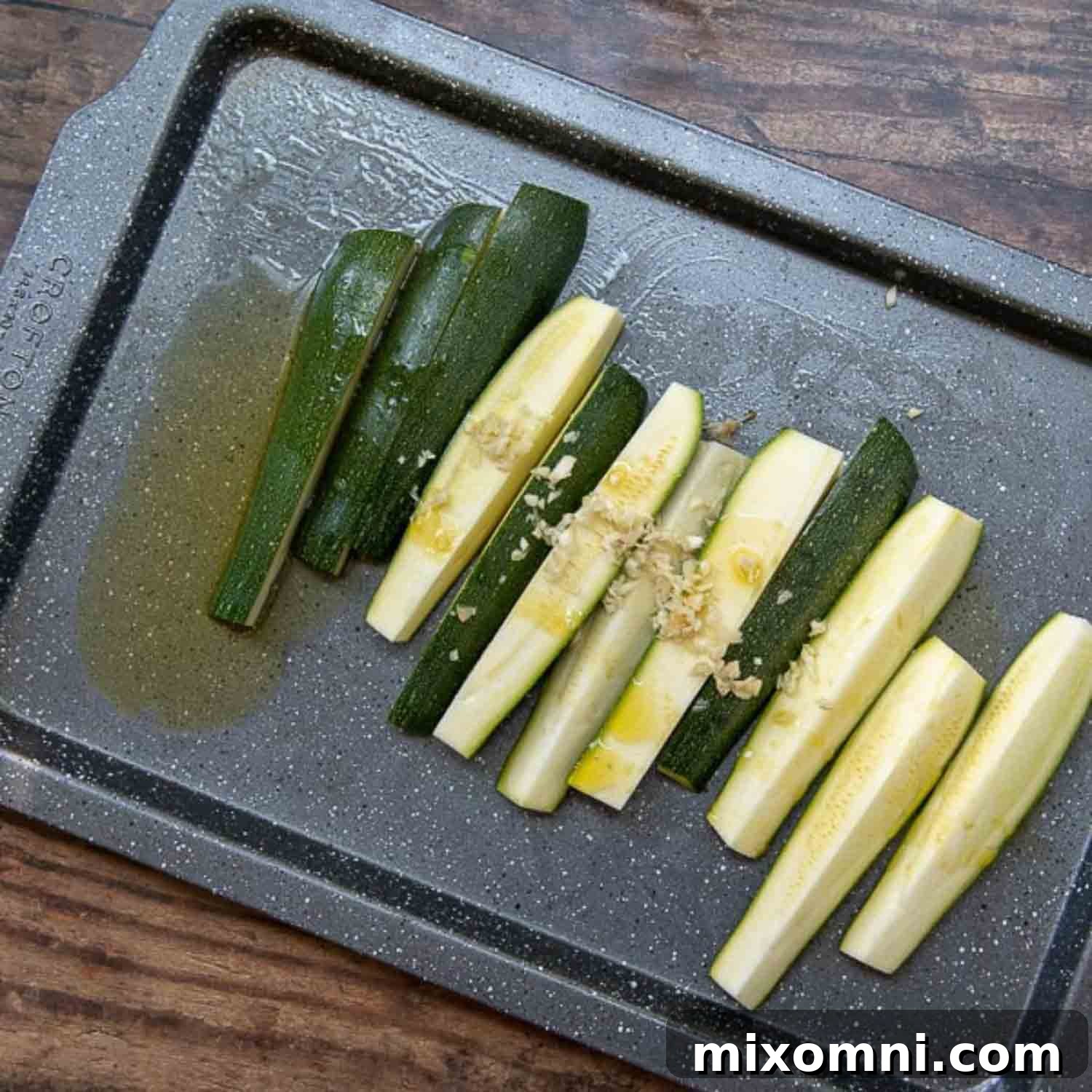 Sliced zucchini spears on a baking sheet, drizzled with olive oil and minced garlic, ready for tossing.