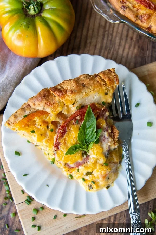 A perfectly sliced piece of tomato pie on a white scalloped plate, with a whole fresh tomato placed beside it.