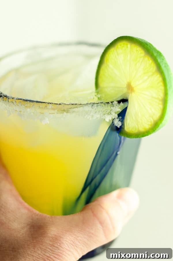 A hand gracefully holding up a single glass of skinny margarita, garnished with lime, against a bright background.
