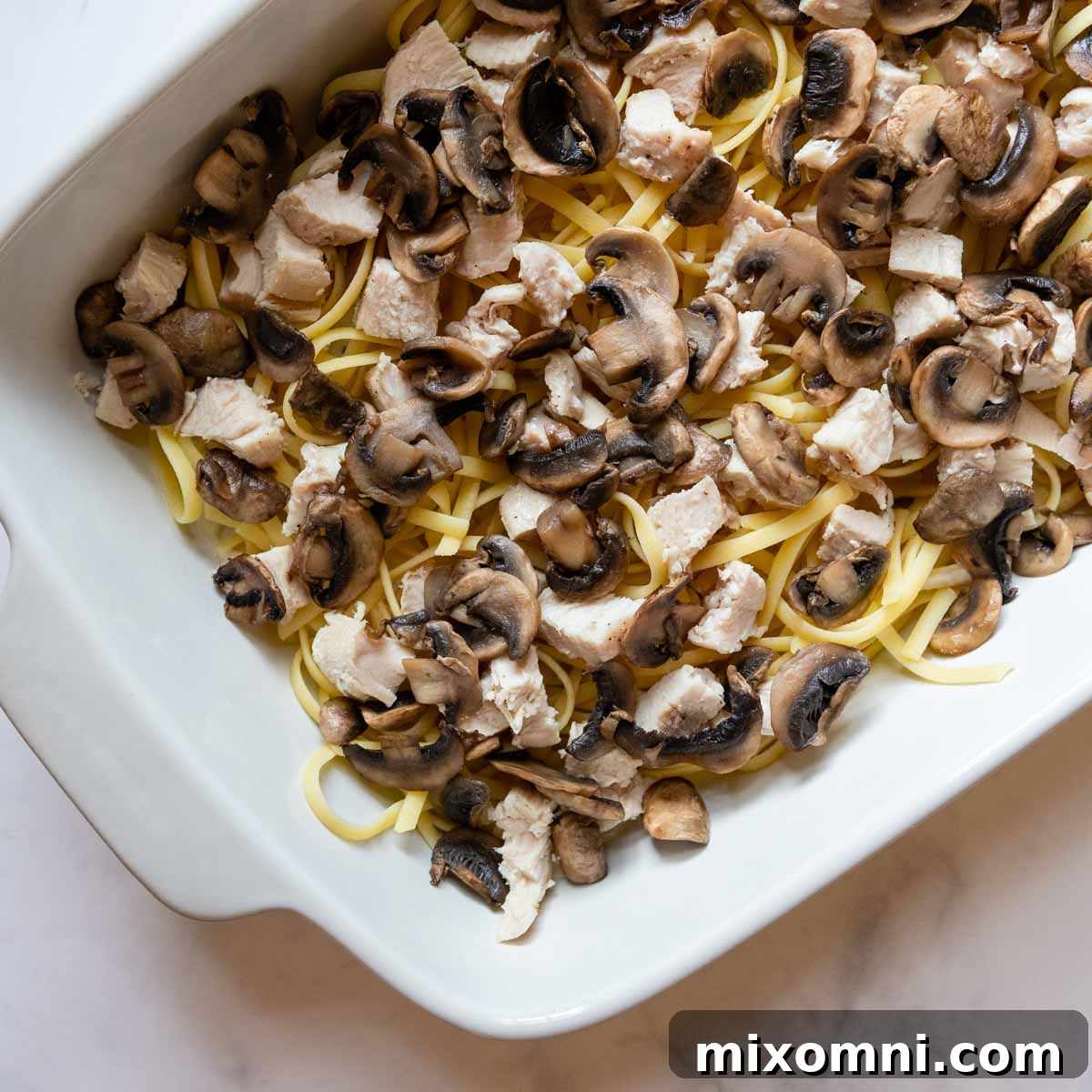 Chopped chicken, mushrooms, and partially cooked noodles layered in a casserole dish.