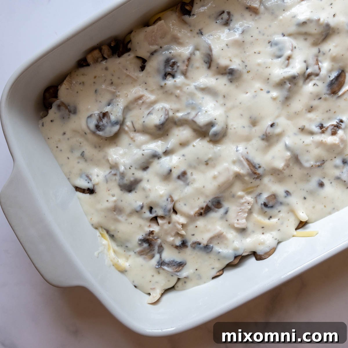 Creamy white sauce poured over the layered mushrooms and chicken in a casserole dish.