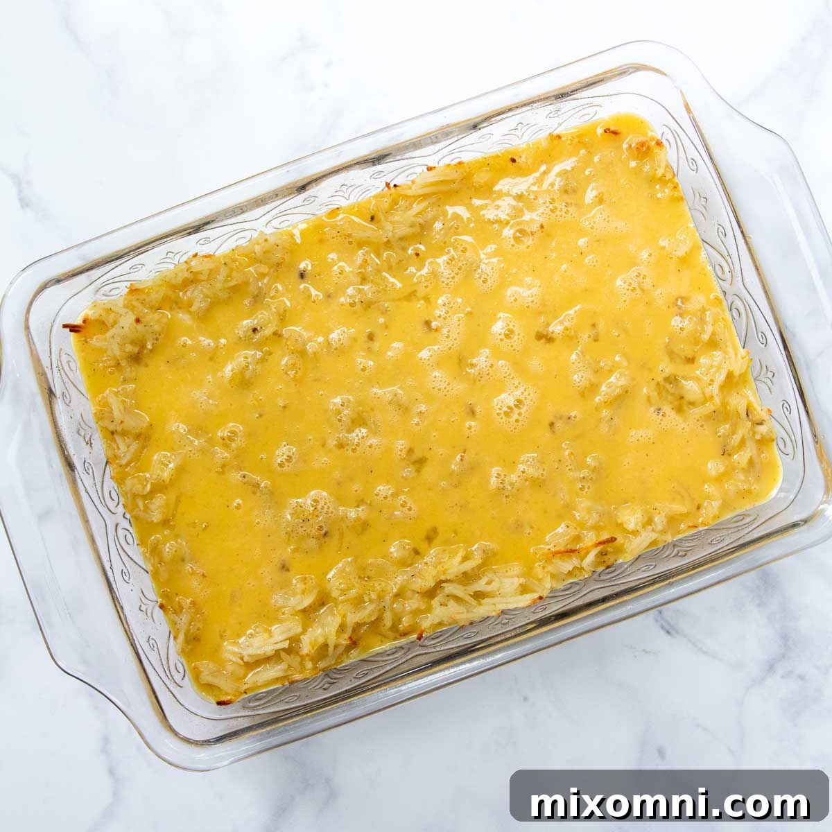 A whisked egg and milk mixture being poured evenly over the golden-brown hash brown layer in a baking dish.