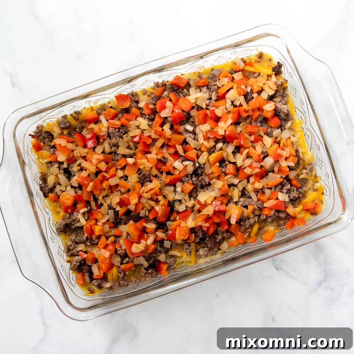 An unbaked breakfast casserole, layered with hash browns, egg mixture, cooked sausage, sautéed vegetables, and a generous sprinkle of cheddar cheese.