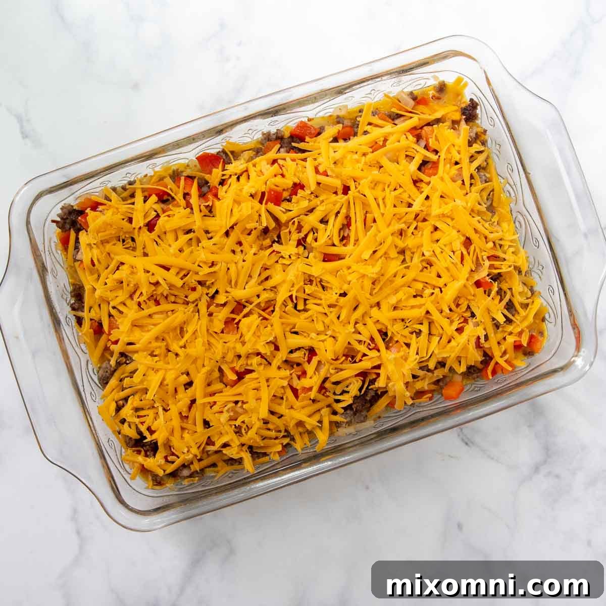 An unbaked breakfast casserole with hash browns, sausage, and vegetables, topped generously with shredded cheese, ready for the oven.