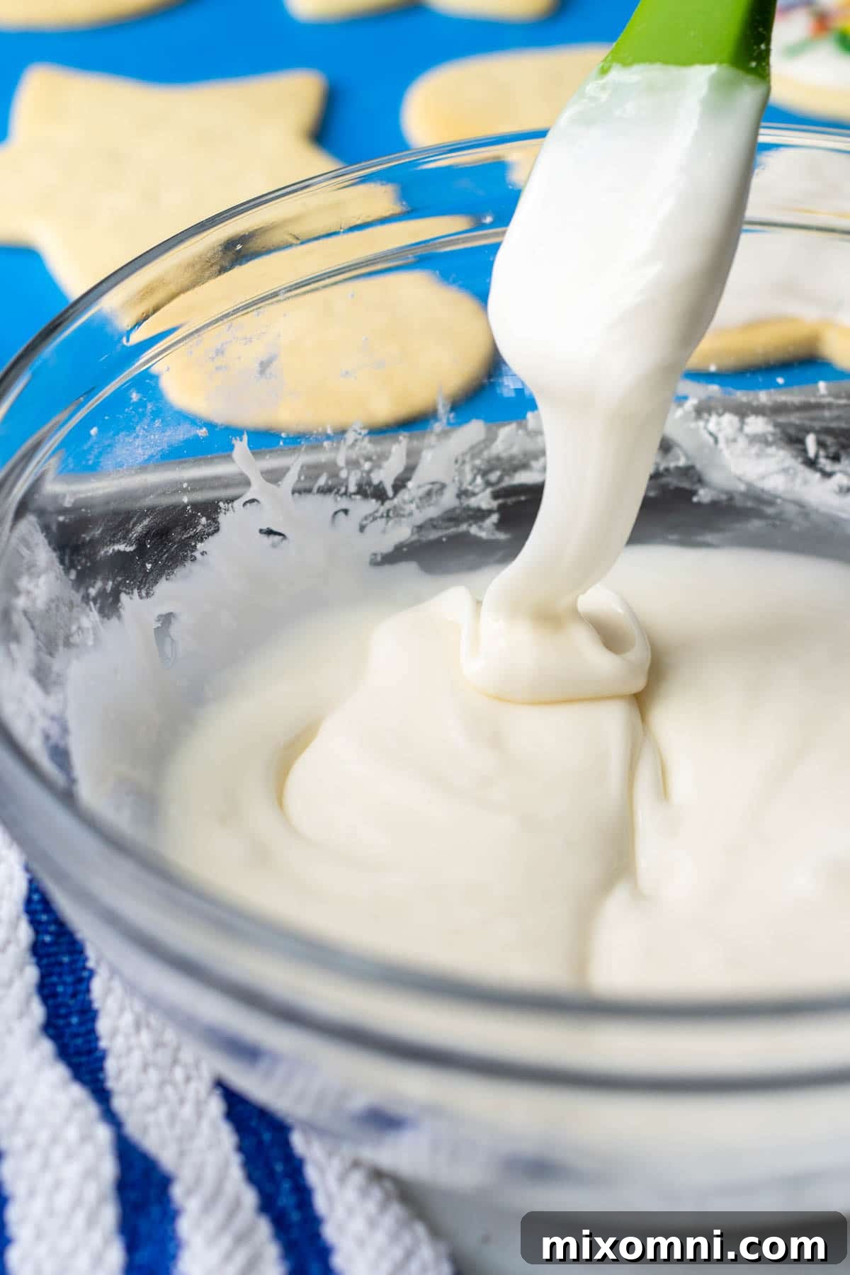 A spoon lifting out creamy, glossy sugar cookie icing from a clear glass bowl, showcasing its perfect smooth texture and readiness for decorating.