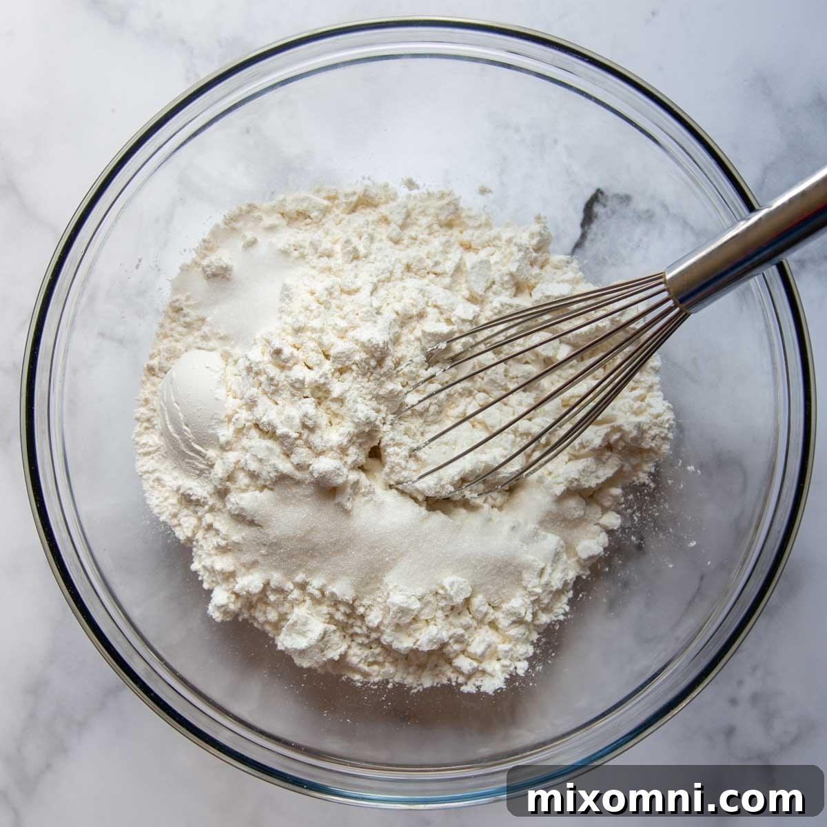 A large mixing bowl containing flour, baking powder, and salt, with a whisk resting inside, ready for the next step.