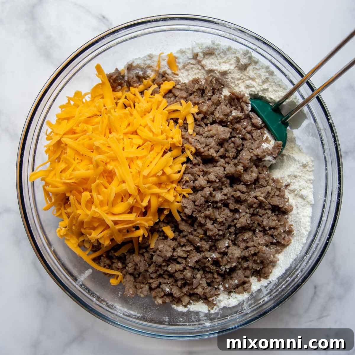 Cooked crumbled sausage and shredded cheddar cheese being added to the dry muffin mixture in a large mixing bowl.