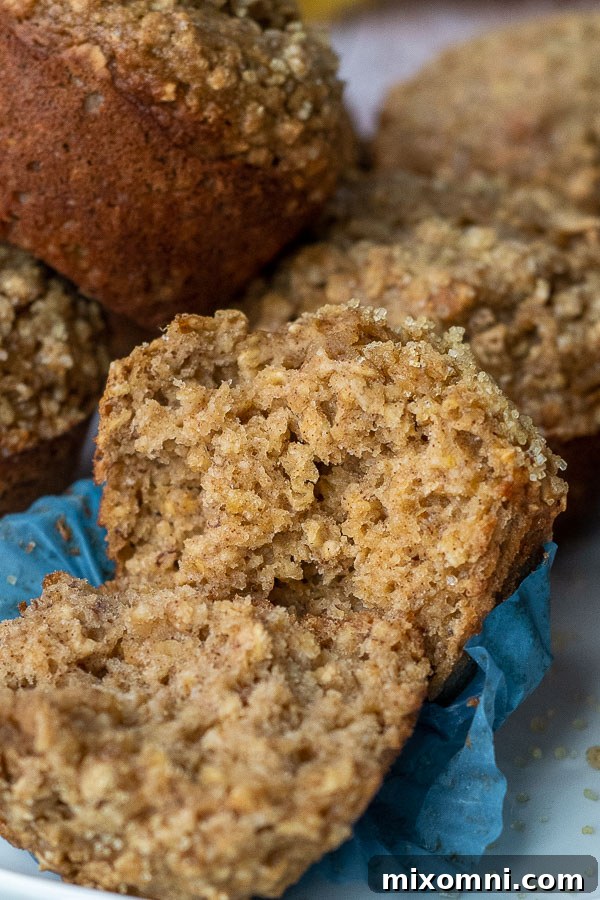 An applesauce oatmeal muffin carefully broken in half, showing its moist, tender interior and hearty oats, surrounded by other wholesome muffins.
