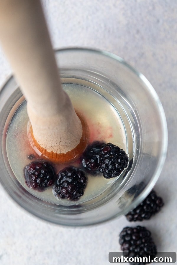 An overhead shot showing fresh blackberries being gently muddled in a glass with a muddler.