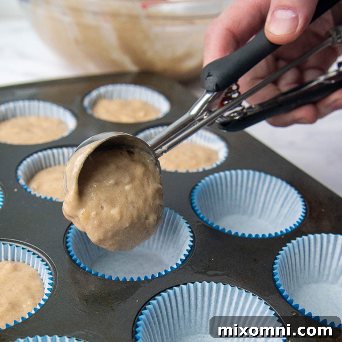 Muffin batter being scooped into paper liners in a muffin tin.