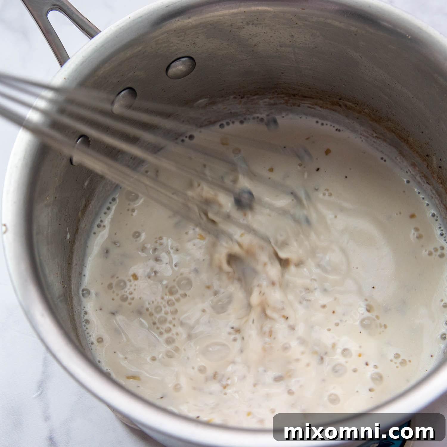 A creamy white sauce being continuously whisked in a saucepan on the stove, showing the development of the béchamel base.