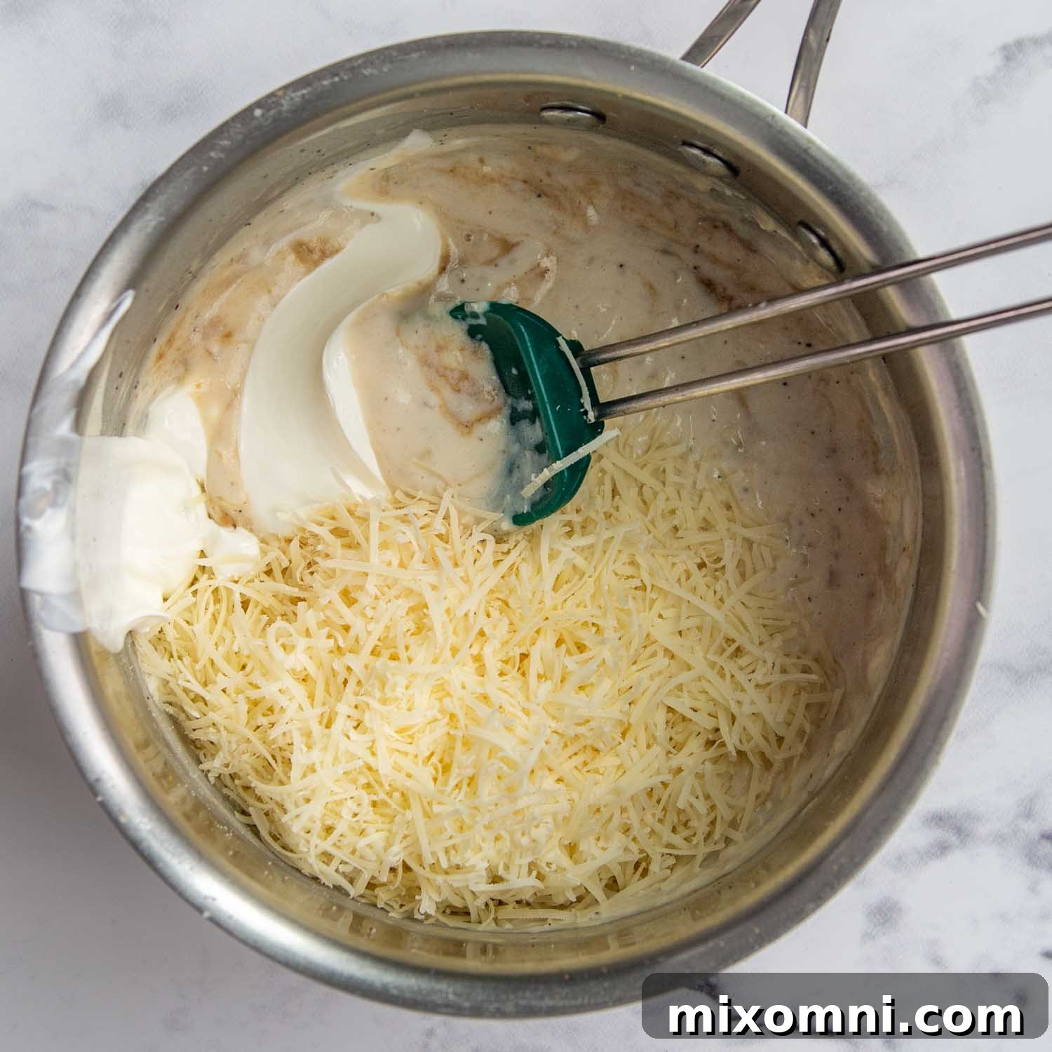A hand stirring freshly grated parmesan cheese and creamy sour cream into the béchamel sauce.