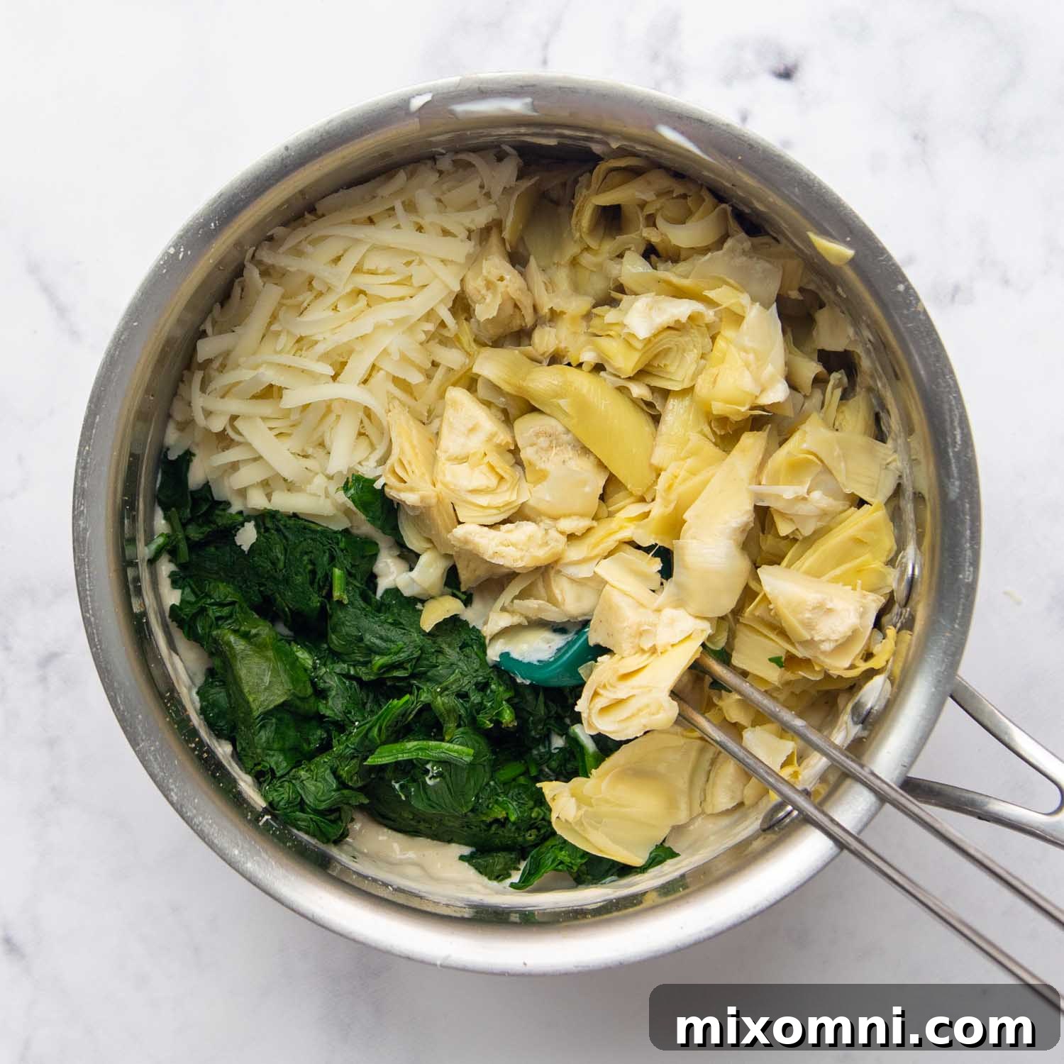 The final steps of adding chopped artichokes and spinach to the rich, cheesy dip mixture in a saucepan.