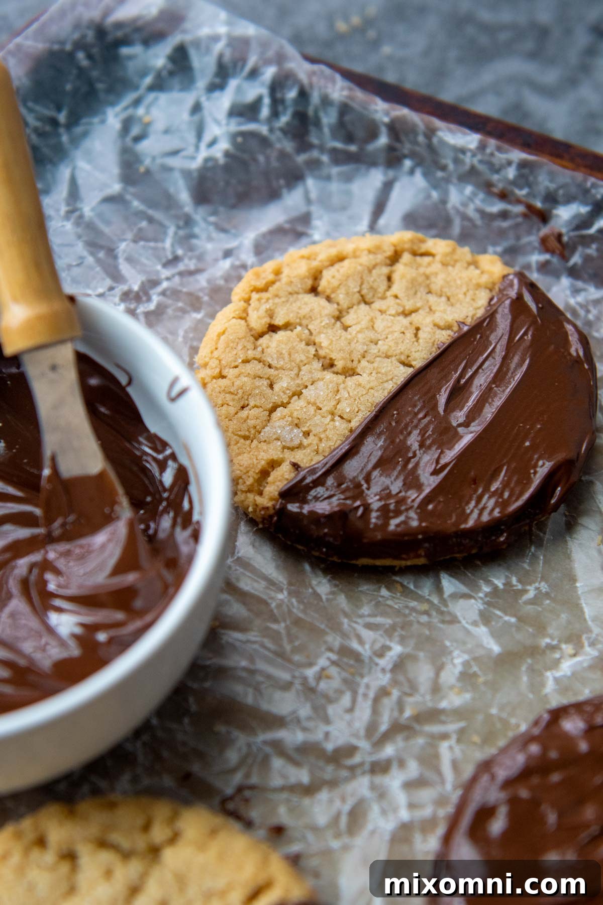 A rich chocolate-dipped almond flour peanut butter cookie sits beside a bowl of melted chocolate, ready for dipping.