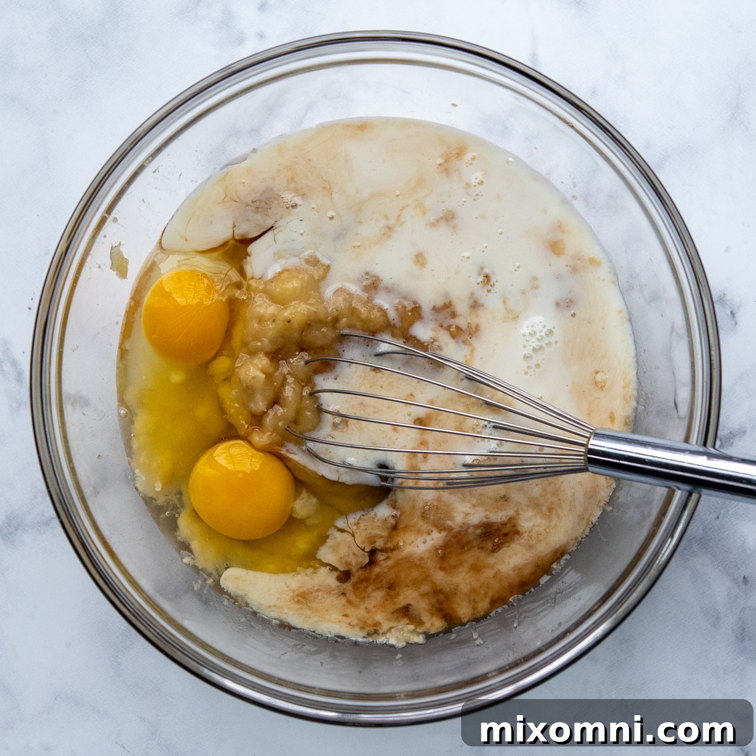 Mashed ripe bananas being combined with milk, eggs, maple syrup, and vanilla extract in a bowl.