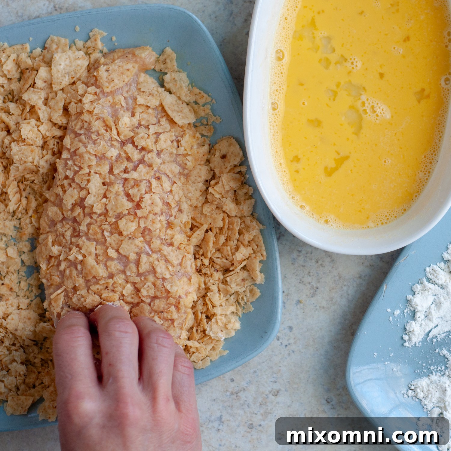 A chicken cutlet being pressed into finely crushed tortilla chips to create a crispy breading.