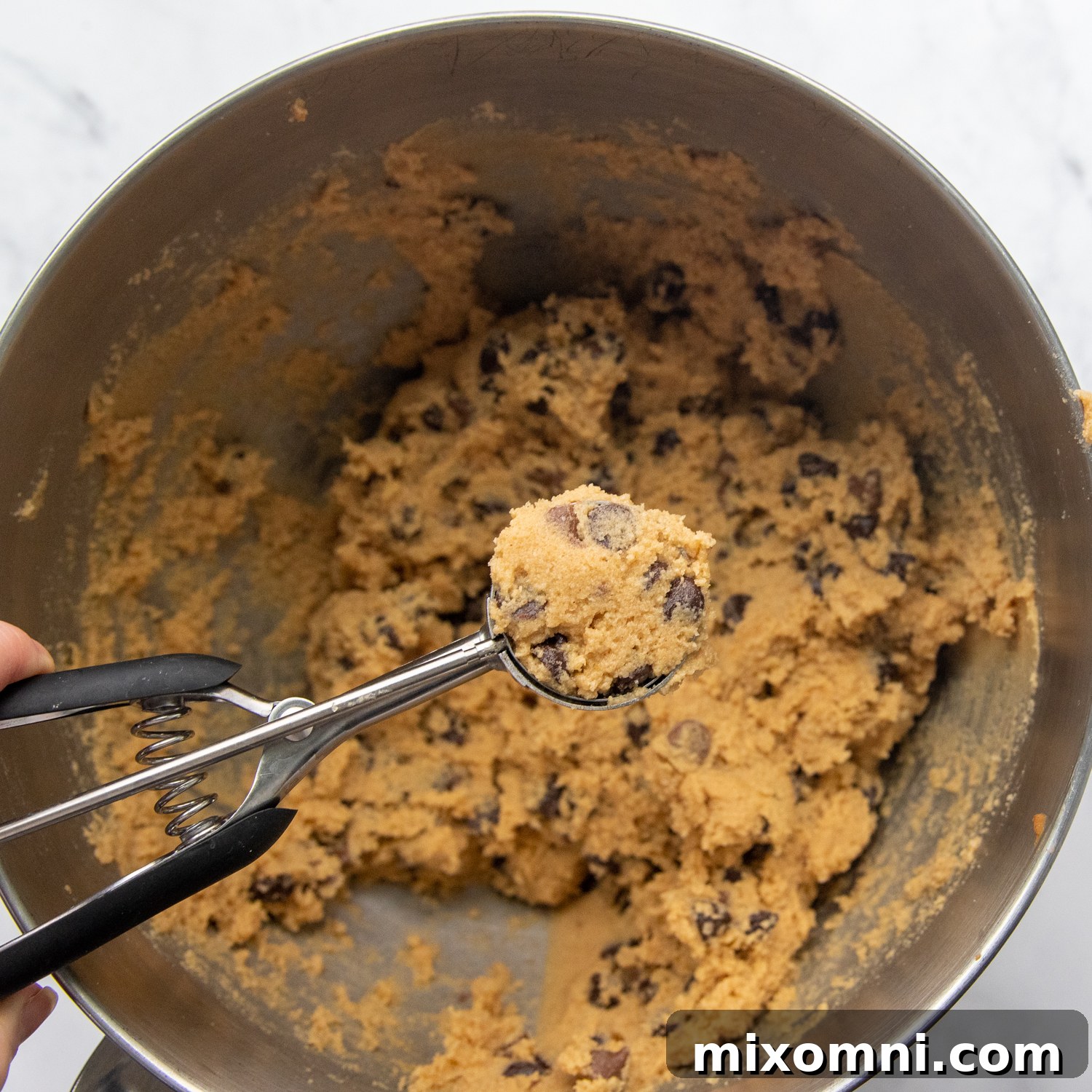A cookie scoop held above a bowl of chocolate chip cookie dough, ready to portion out perfect cookies.
