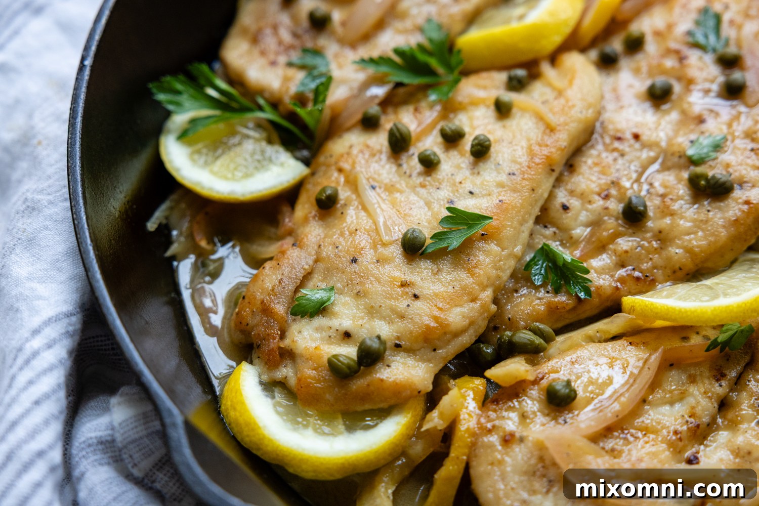 Chicken cutlet in a skillet with lemon garnish and topped with capers.